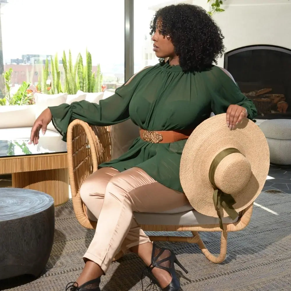 A glamorous model sits on a chair holding the satin lined raffia wide brimmed straw hat with the 100% silk olive green bow.