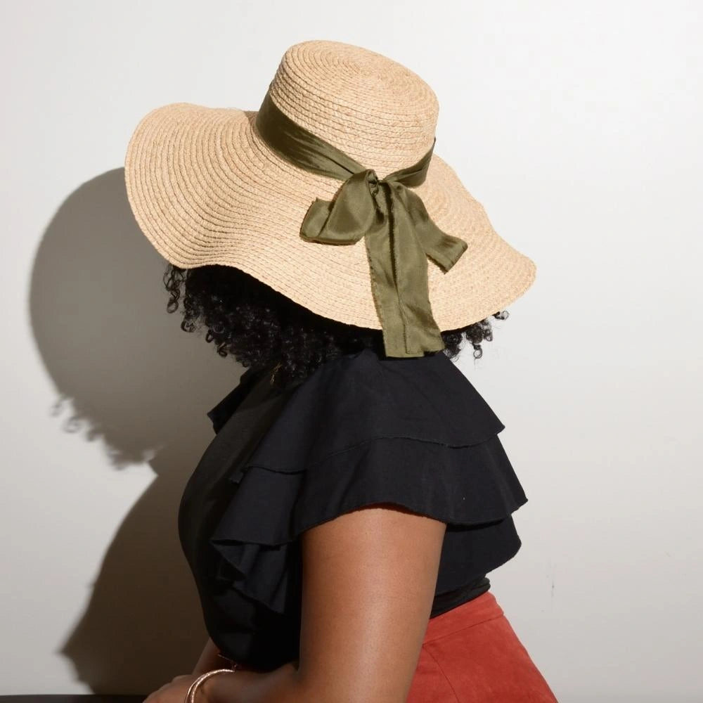 The back of a model wearing the satin lined raffia wide brimmed straw hat with the 100% silk olive green bow.